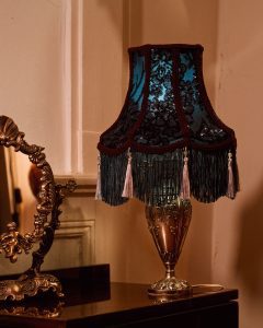 Géraldine: Lampe de table artisanale avec abat-jour en dentelle noire sur fond bleu, orné de franges et pompons, à côté d'un miroir doré ciselé.