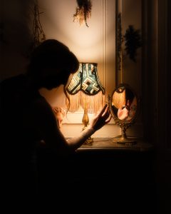 Lampe de table artisanale au style Art Déco avec abat-jour à motifs géométriques et franges, reflétée dans un miroir ovale doré.